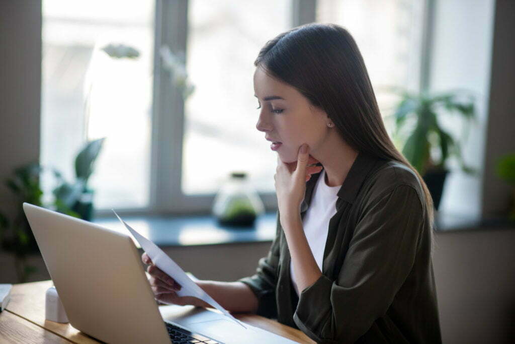 Girl sitting in front of laptop carefully looking at paper document. aca