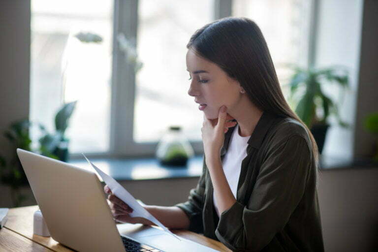 Girl sitting in front of laptop carefully looking at paper document. aca