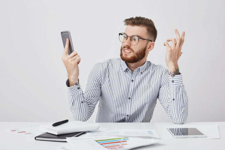 Angry businessman with trendy hairstyle, wears round spectacles, looks furiously into cell phone, ca
